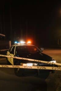 A police car with emergency lights at a night crime scene, surrounded by yellow police tape.