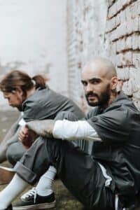 Two inmates with tattoos sitting against a brick wall wearing prison uniforms. Intense expressions.