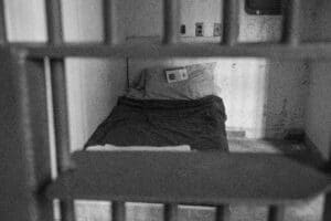 Black and white photo of an empty prison cell with a bed and book.