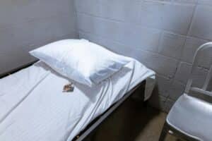 A stark prison cell with a single bed, white linens, a pillow, and a chair offering a minimalist view.