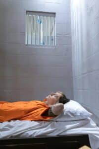 A woman in orange prison attire lies on a bed in a stark jail cell.