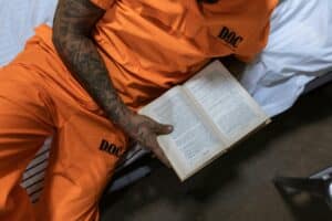 Tattooed inmate reading a book on a bed in a prison cell.