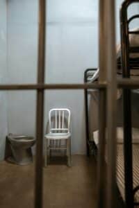Empty prison cell with metallic furniture including bunk bed, chair, and toilet in a confined space.