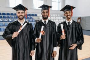 Three young men in graduation gowns and mortarboards smiling and holding diplomas indoors.