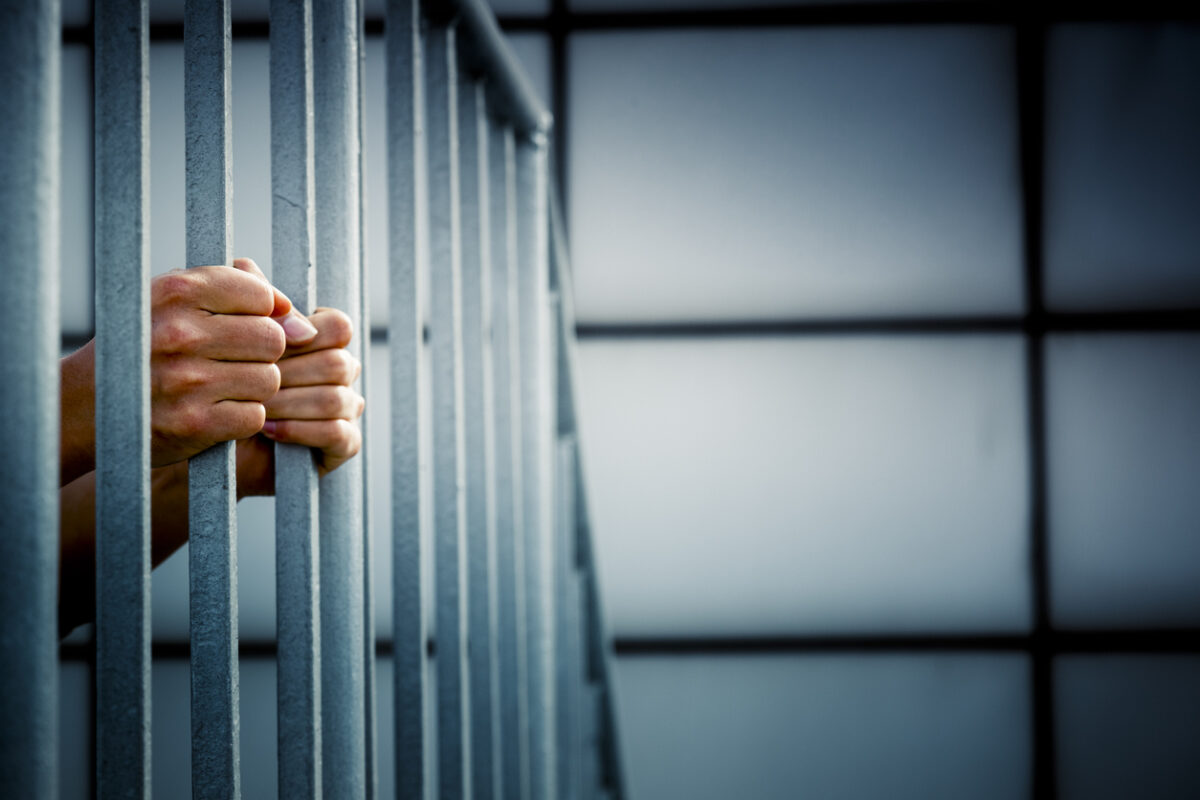 A hand gripping prison bars in a darkened cell.
