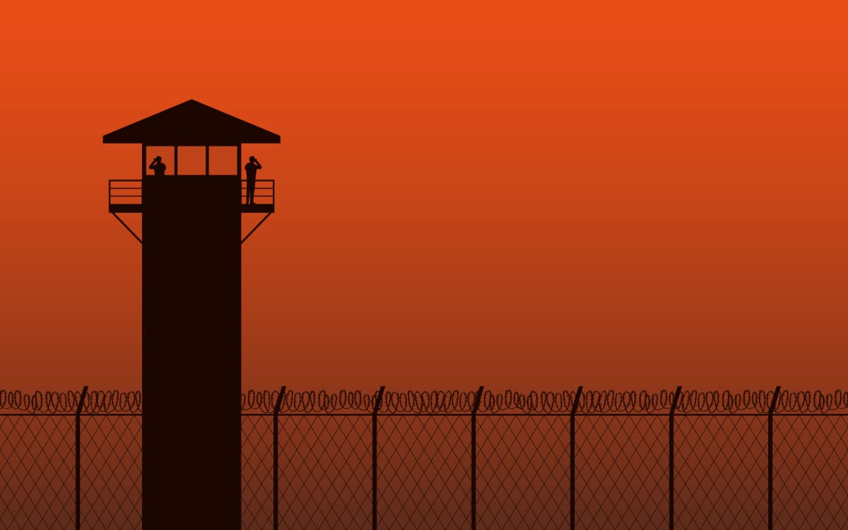 A guard tower silhouetted against an orange sky above a chain-link fence.