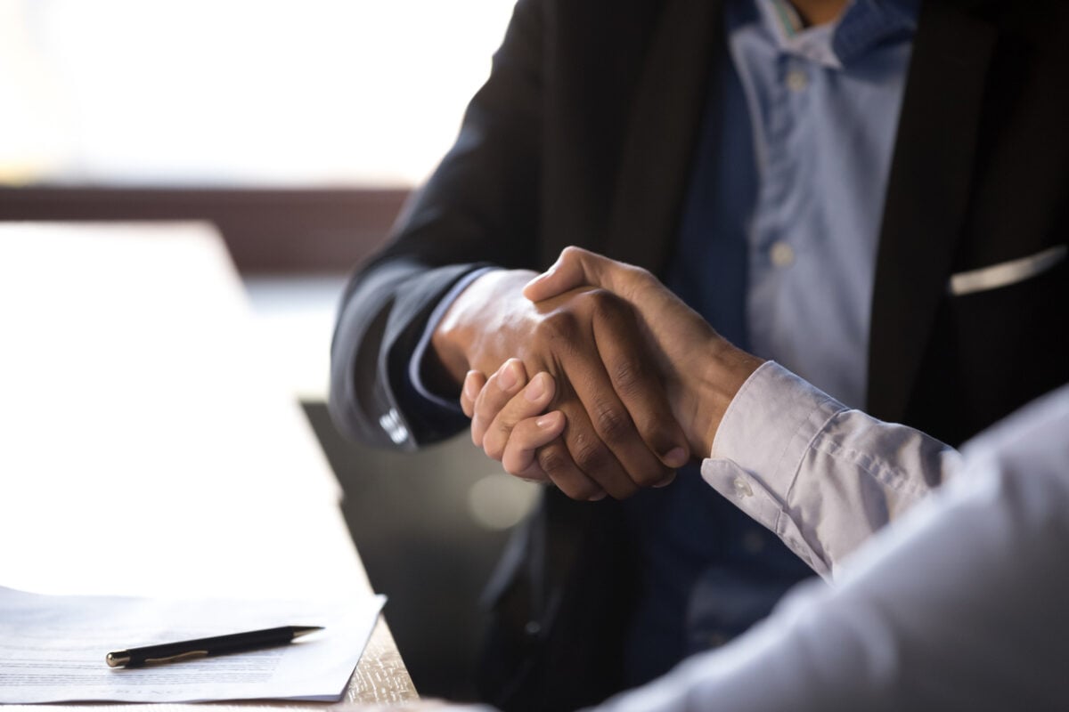 Two people in suits shaking hands across a desk.