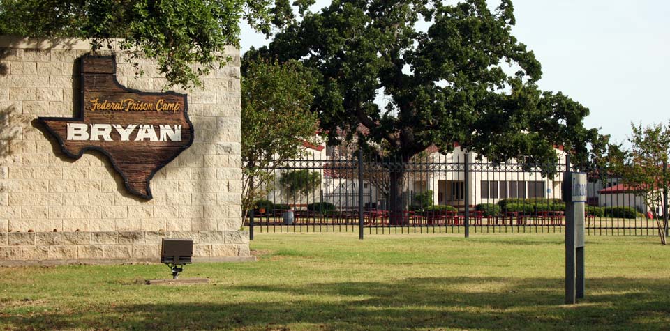 FPC Bryan FPC Bryan, Texas-shaped Federal Prison Camp sign on a stone wall, with an oak and white buildings behind an iron fence